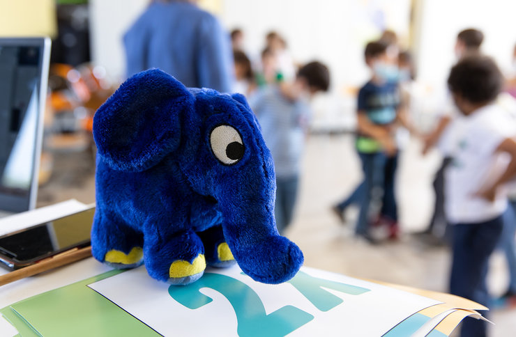 Der blaue Elefant als Plüschtier auf Papieren, im Hintergrund viele Kinder unscharf