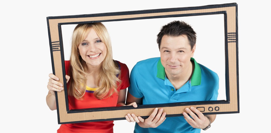 Die Moderator:innen Tanja und André schauen durch einen TV-Rahmen aus Pappe