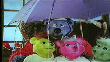 Käptn Blaubär steht mit seinen Enkeln unter einem Regenschirm.