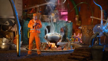 Mann in orangefarbener Arbeitskleidung steht on einer Fabrikhalle vor einem dampfenden Kessel