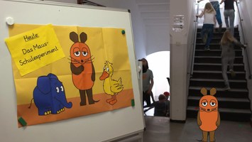 Maus steht in einem Schultreppenhaus vor einem Aufsteller mit einem Mausplakat 