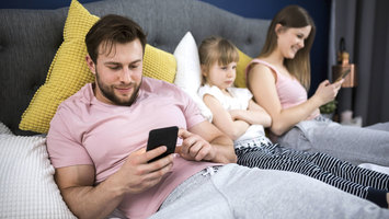 Mädchen liegt zwischen ihren Eltern auf dem Bett und ist sauer, dass beide Eltern sich mit ihren Handys beschäftigen