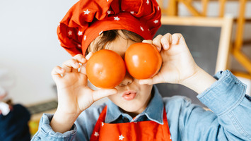 Junge in Kochmütze und Schürze hält sich Tomaten vor die Augen