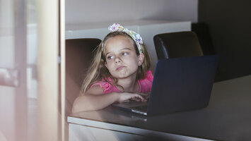 Ein Mädchen sitzt an ihrem Laptop und schaut traurig nach draußen.