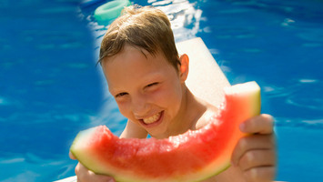 Ein Junge sitzt am Rand eines Schwimmbeckens und hält eine Melone in die Kamera