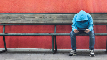  Jugendlicher mit Kapuze sitzt auf einer Bank und schaut ins Tablet