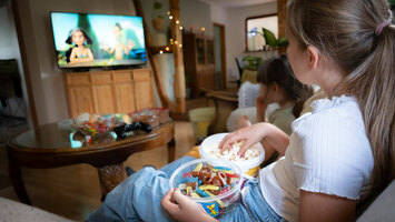 Zwei Kinder sitzen im Wohnzimmer auf dem Sofa und essen Popcorn und Süßigkeiten und schauen dabei Fernsehen.