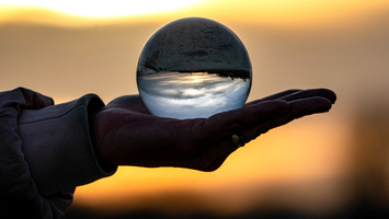 Hand hält eine Glaskugel in der sich die Umgebung und der Sonnenuntergang spiegeln.