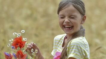 Ein Mädchen sitzt in einem Kornfeld, hält einen Blumenstrauß in der Hand und lacht.