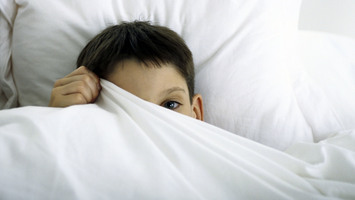 Junge liegt im Bett und zieht sich die Decke fast gänzlich vors Gesicht