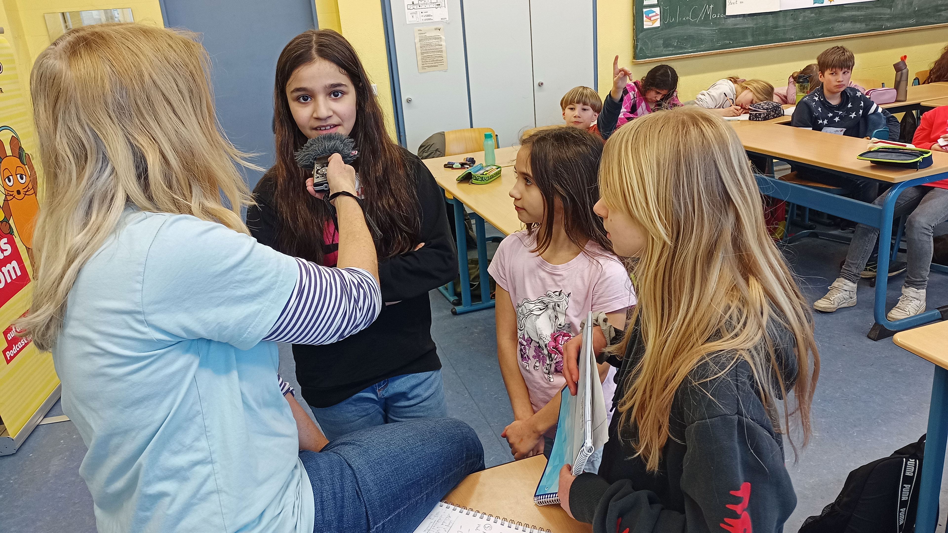 Schülerinnen und Schüler werden in ihrer Klasse von einer Reporterin interviewt.