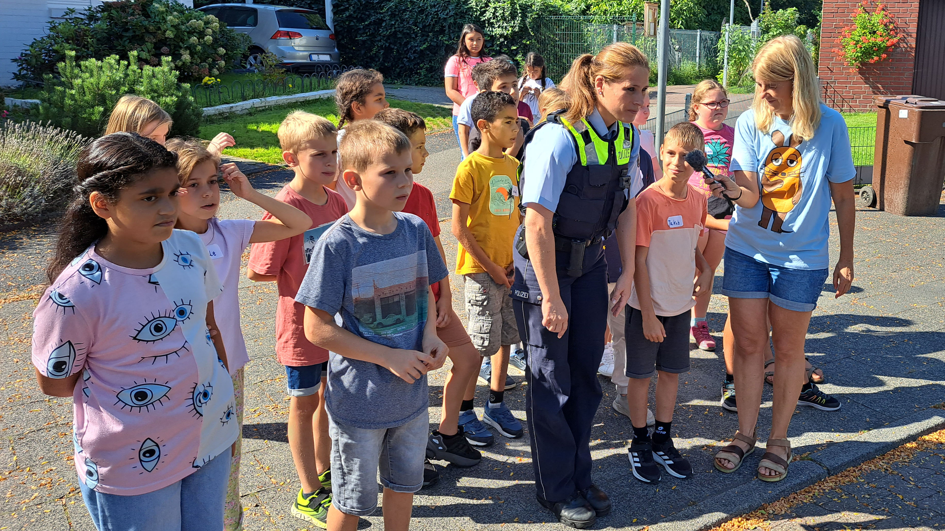 Kinder der MausKlasse stehen mit Polizistin Sandra Schmidt und Mausreporterin Christina Pannhausen auf dem Gehweg