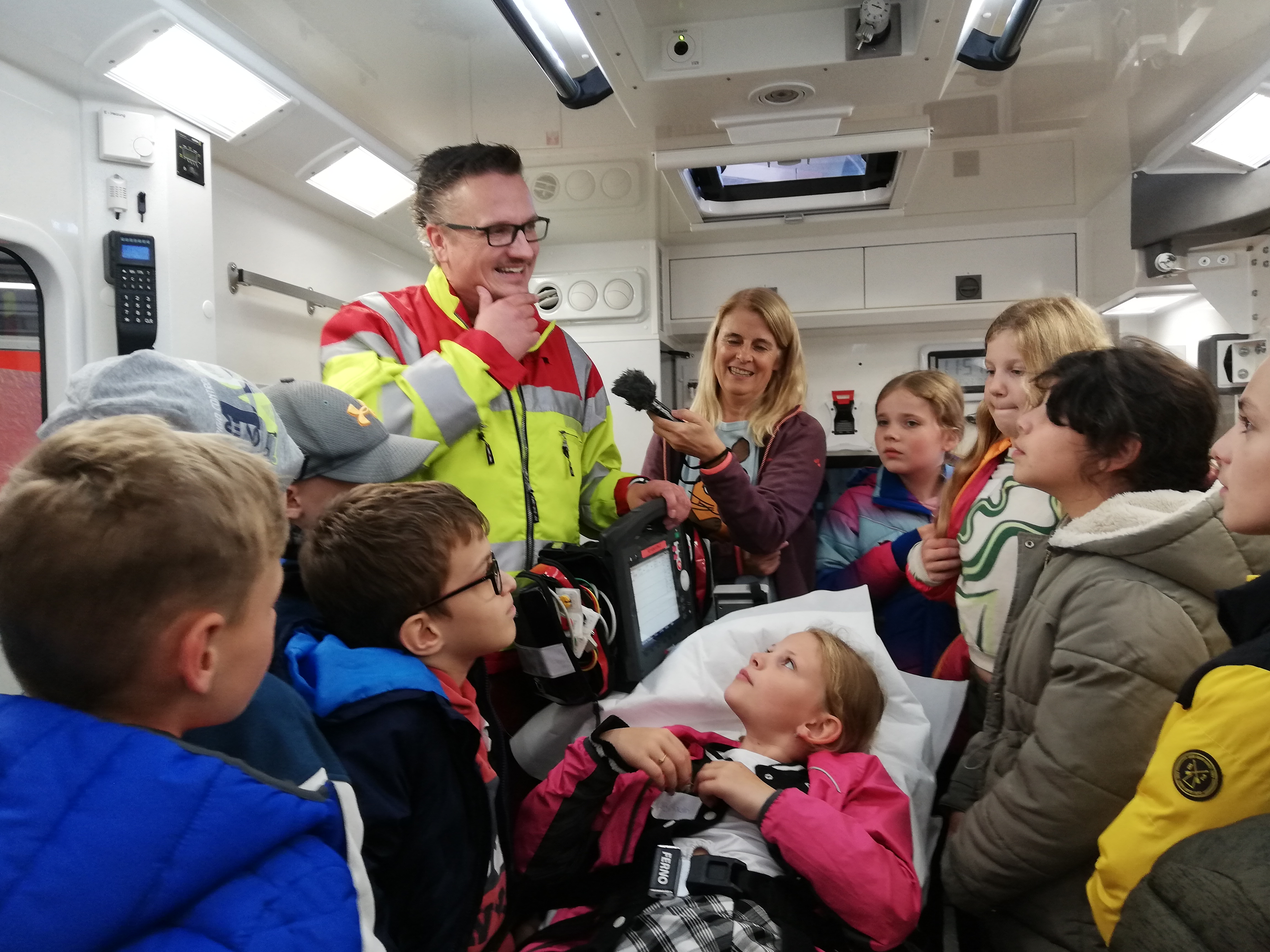 Eine Schulklasse steht in einem Rettungswagen, befragt einen Sanitäter und ein Mädchen liegt auf einer Trage.