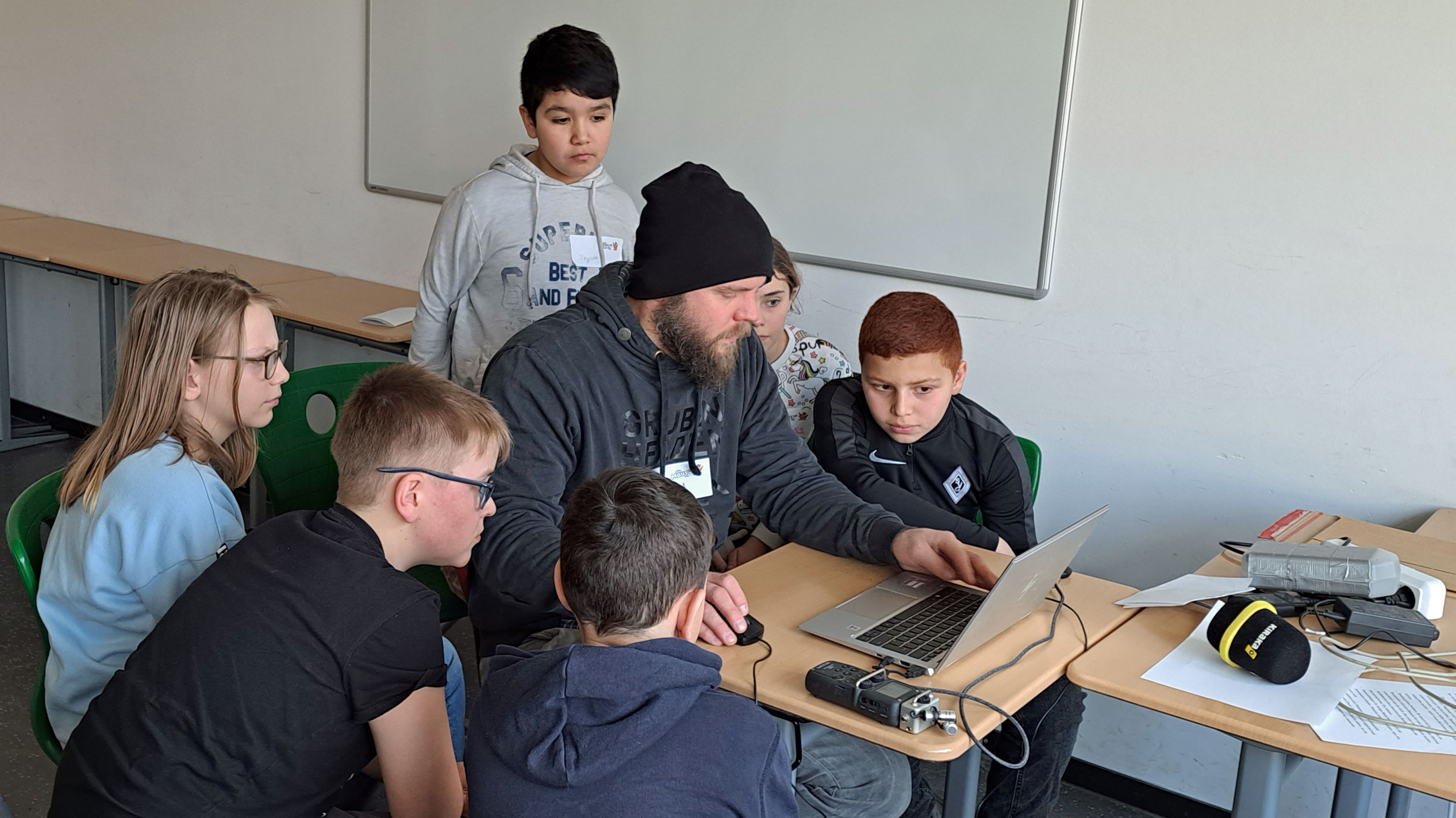 Schülerinnen, Schüler und ein Erwachsener sitzen vor einem Laptop.