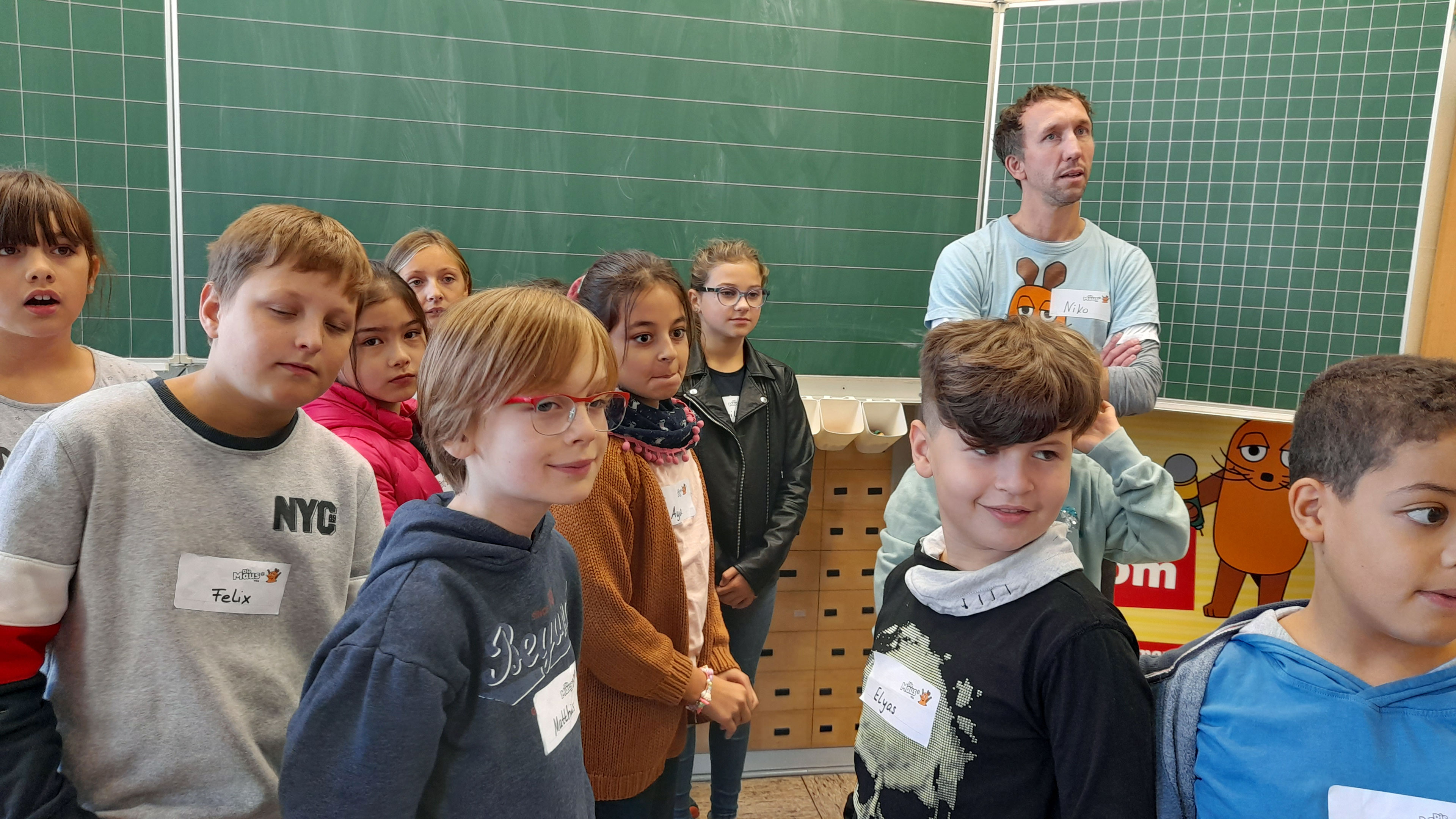 Kinder und ein Mann stehen vor einer Tafel.