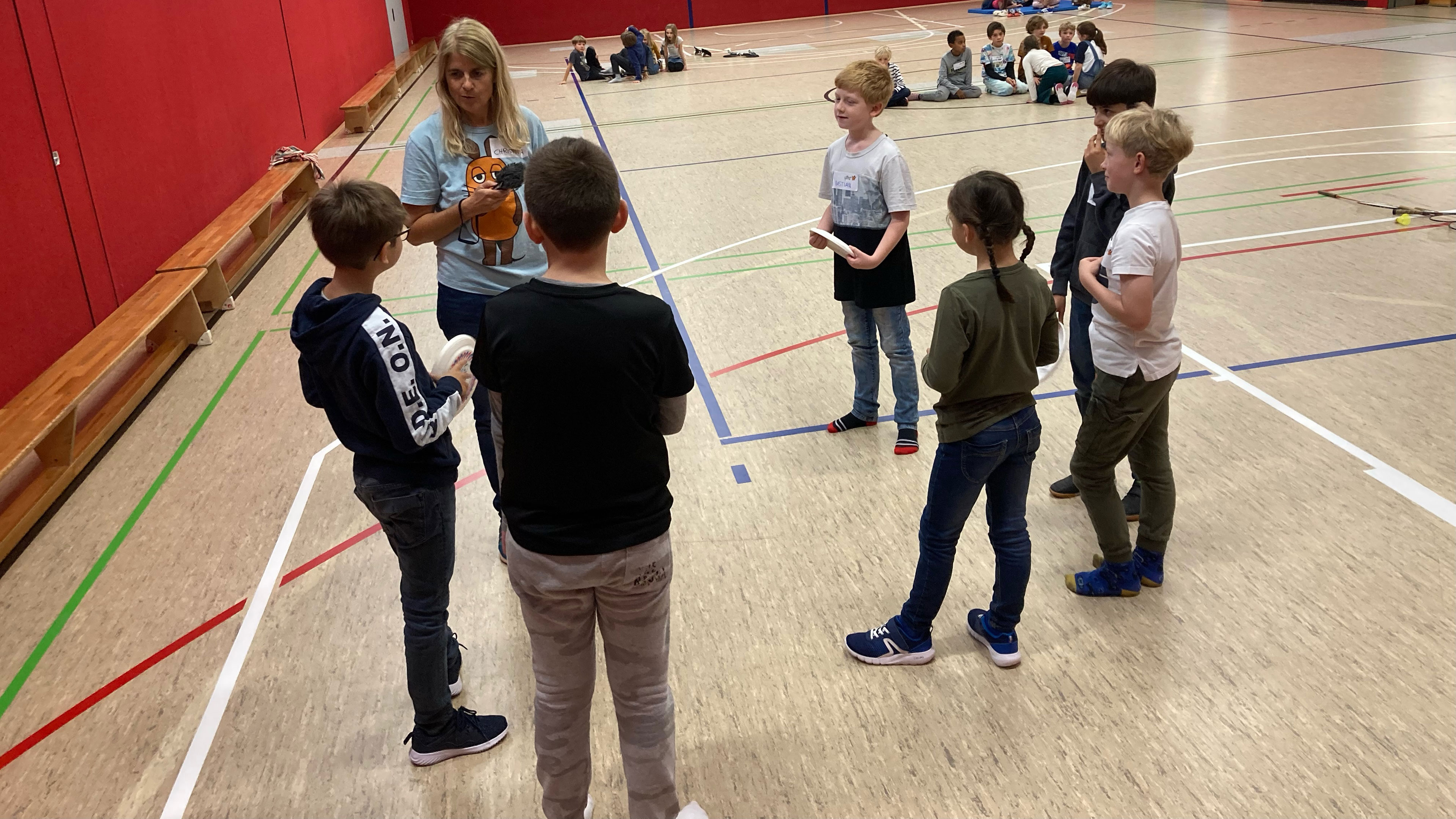 Kinder stehen mit einer Reporterin in einer Turnhalle im Kreis.