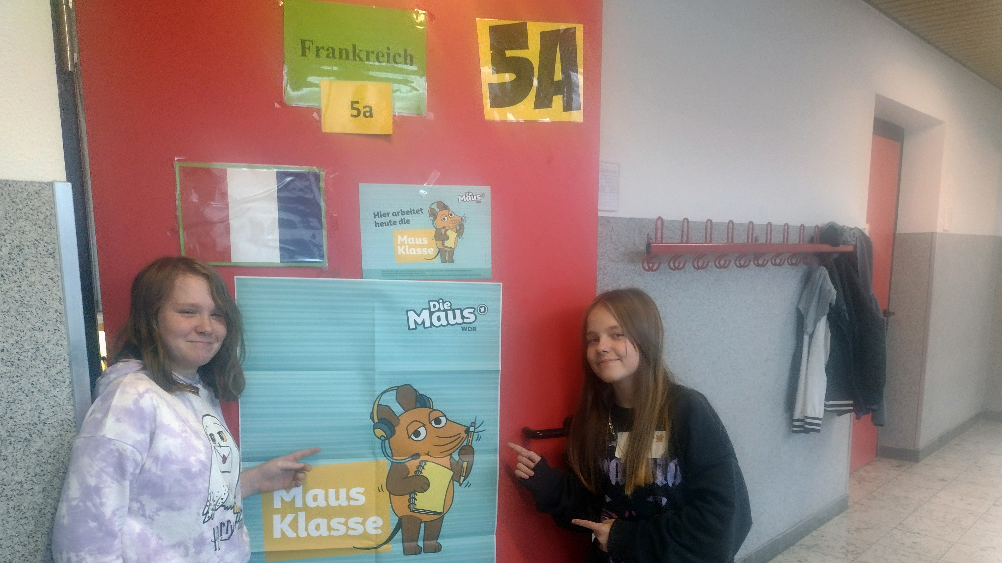 Zwei Mädchen zeigen auf die Tür ihres Klassenzimmers, an die ein Maus-Poster geklebt ist