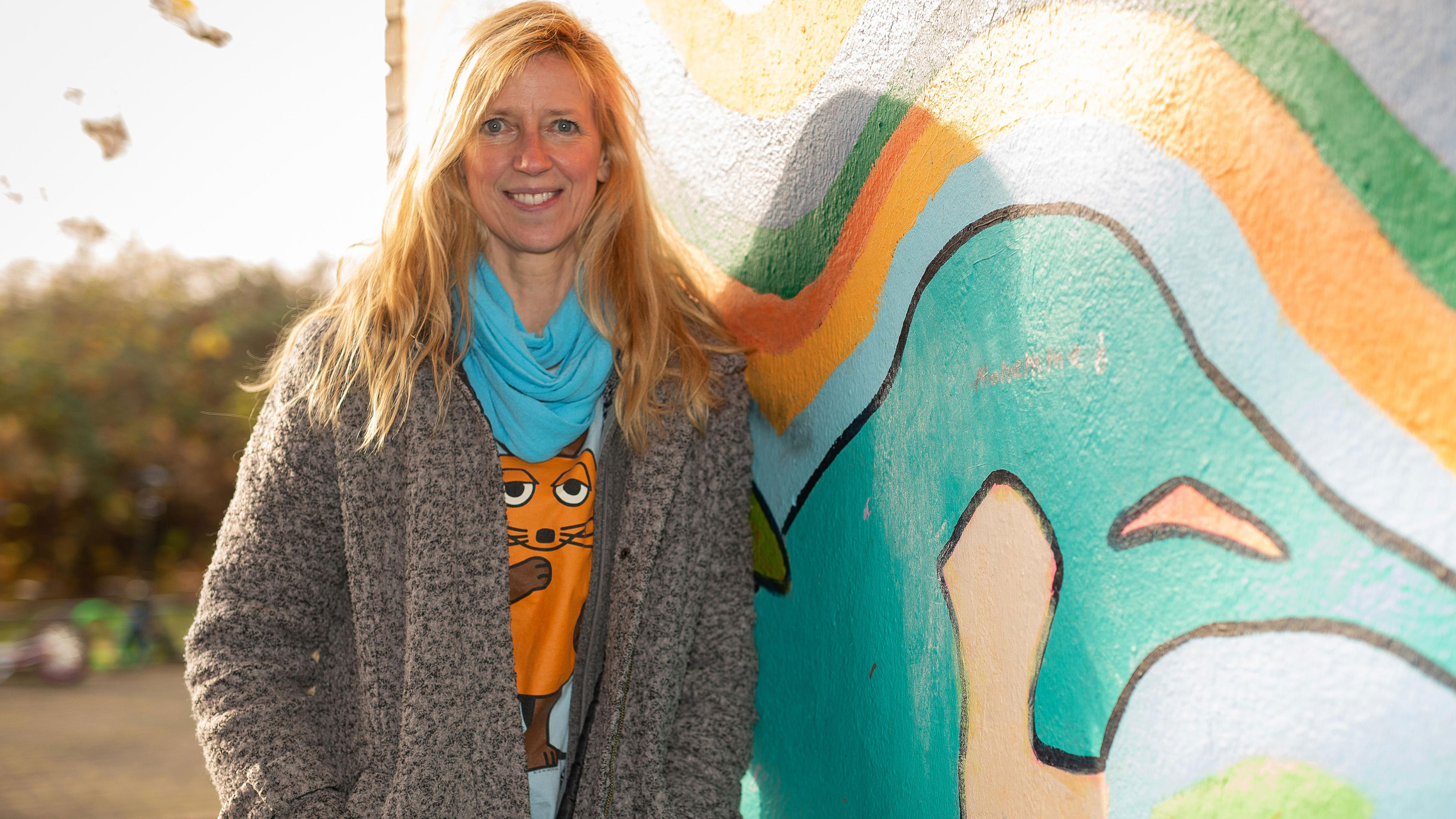 Eine Frau mit langen blonden Haaren, die unter ihrem Mantel ein T-Shirt mit der Maus darauf trägt, lehnt sich an eine mit buntem Graffiti bemalte Hauswand.