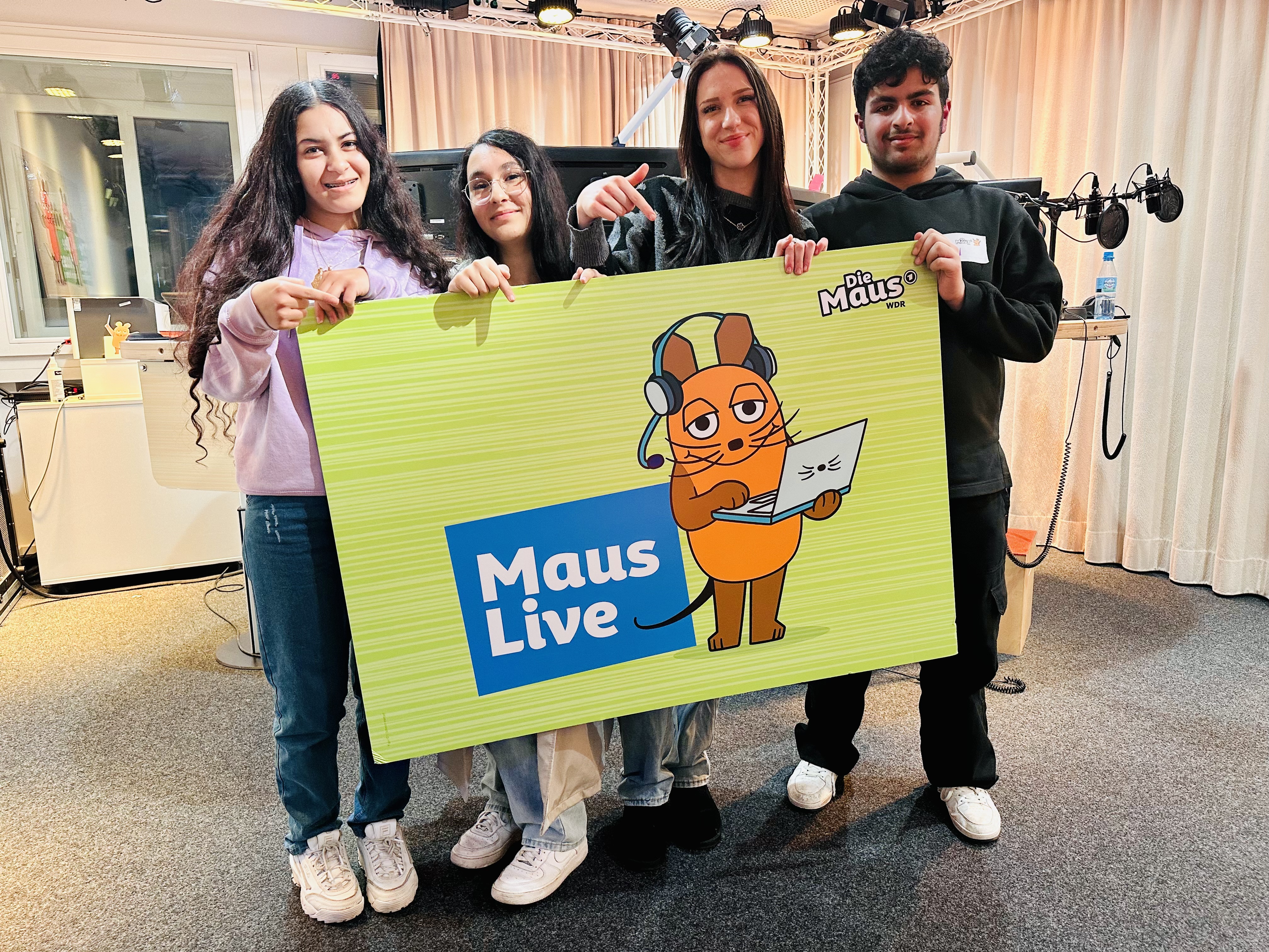 Drei Schülerinnen und ein Schüler halten ein MausLive-Plakat in den Händen.