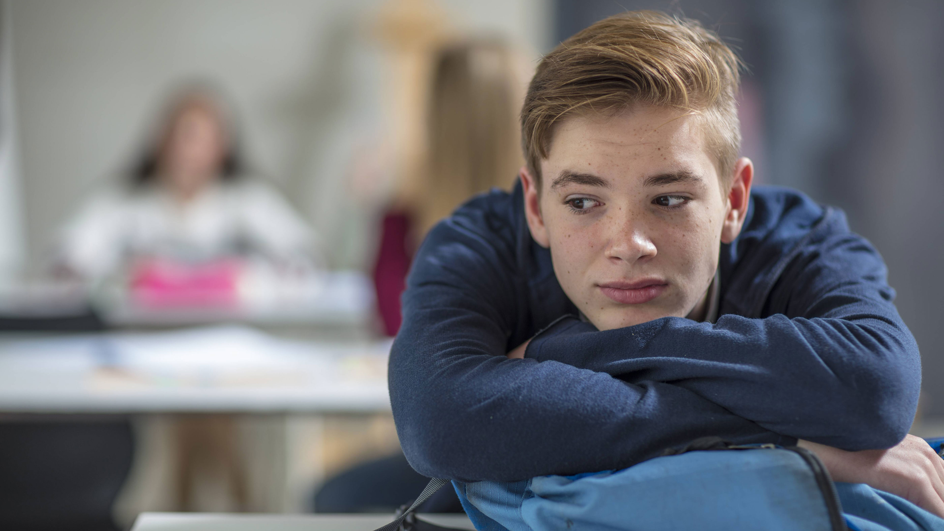 Ein Junge sitzt nachdenklich in einer Klasse und hat sich auf seinen Rucksack gelegt.