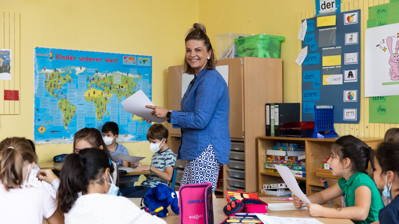 Schulklasse mit Kindern an Tischen, Lehrerin verteilt Zettel