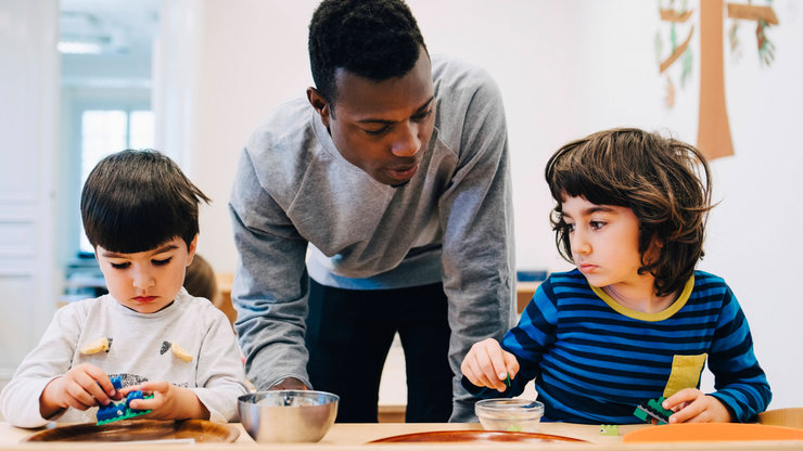 Männlicher Lehrer arbeitet mit Kindern am Tisch und erklärt ihnen etwas.