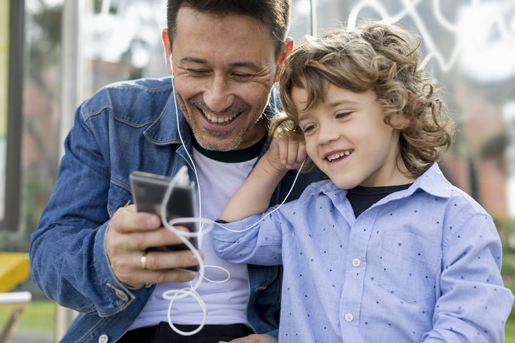 Ein lachender Mann und ein Junge teilen sich Kopfhörer und schauen dabei auf ein Smartphone