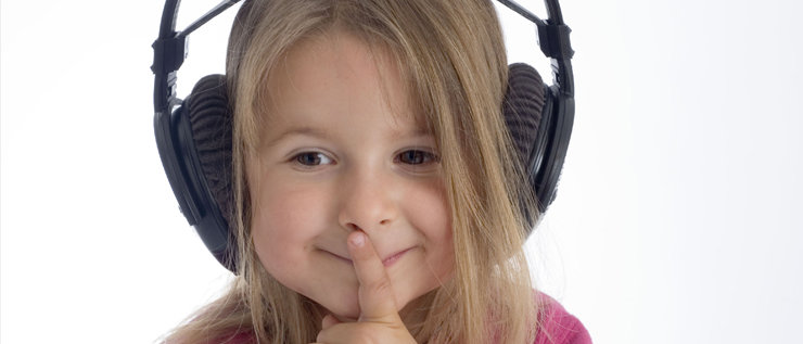 Ein kleines Mädchen mit großen schwarzen Kopfhörern lauscht und hat den Zeigefinger über die Lippen gelegt