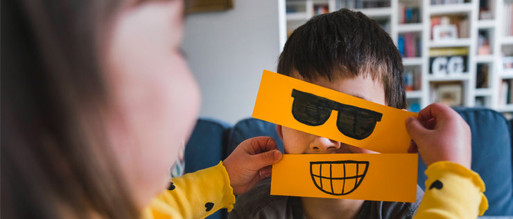 Ein Kind hält einem anderen eine auf orangefarbenes Papier gemalte Sonnenbrille und einen Mund vor das Gesicht