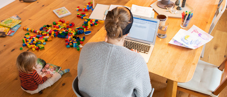 Mutter mit Laptop am Tisch und ein Kind sitzt auf dem Boden und liest ein Buch.