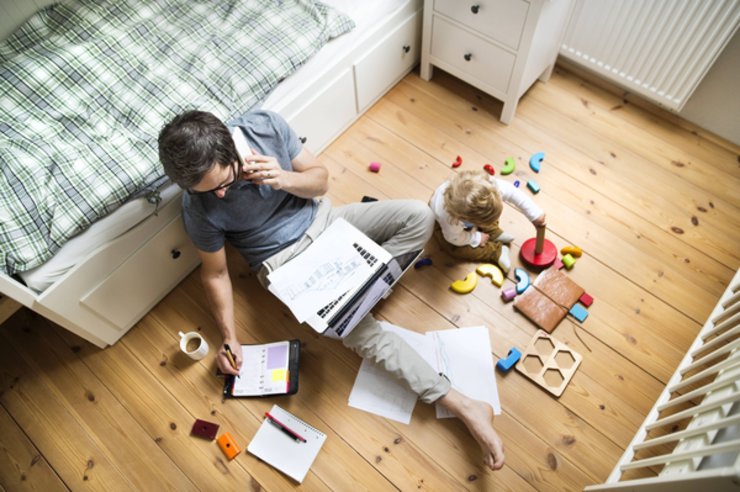 Vater mit Kind sitzt auf dem Boden. Er arbeitet am Laptop und das Kind spielt.