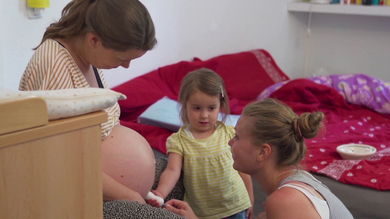 Schwangere Frau wird von kleinem Mädchen mit dem Dopton untersucht, Hebamme sitzt daneben