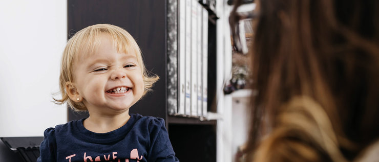 Mutter sitzt am Schreibtisch vor dem Computer und das Kind neben ihr auf dem Schreibtisch