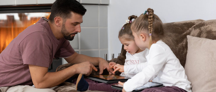 Vater sitzt vor ein Couch, auf der Zwillinge mit einem Tablet sitzen.