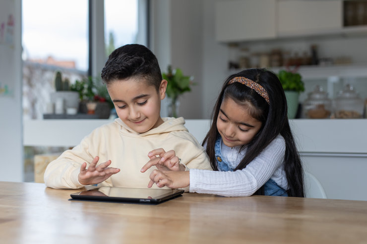 Ein Junge und ein Mädchen sitzen an einem Tisch und tippen auf ein Tablet.