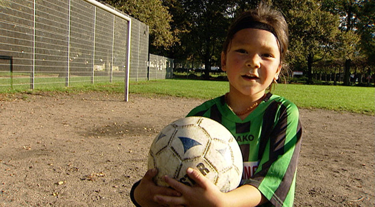 Ein Junge mit Stirnband hält draußen einen Fußball fest. Bild aus der Sachgeschichte Kelana spielt Fussball