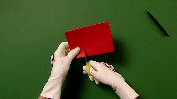 Zwei Hände mit Handschuhen schneiden Schlitze in das rote Papier.