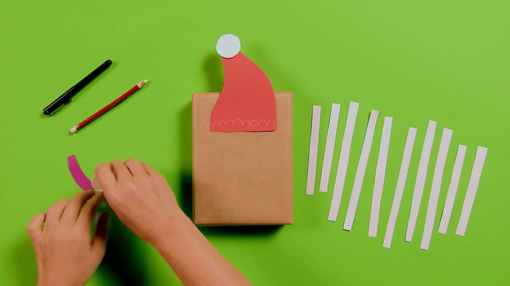 Zwei Hände nehmen Papierteile zum Paket mit aufgeklebter roter Mütze auf grünem Grund