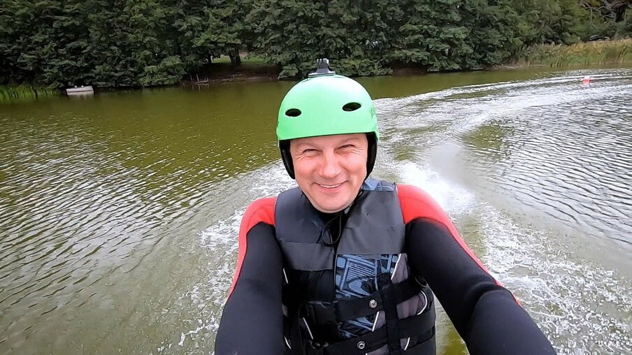 Jonathan fährt mit einem Neoprenanzug und einem Helm auf einer Wasserskianlage