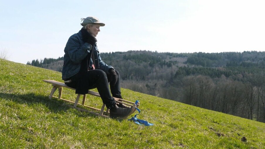 Laura sitzt auf einem Schlitten auf auf einem grünen Wiesenhang